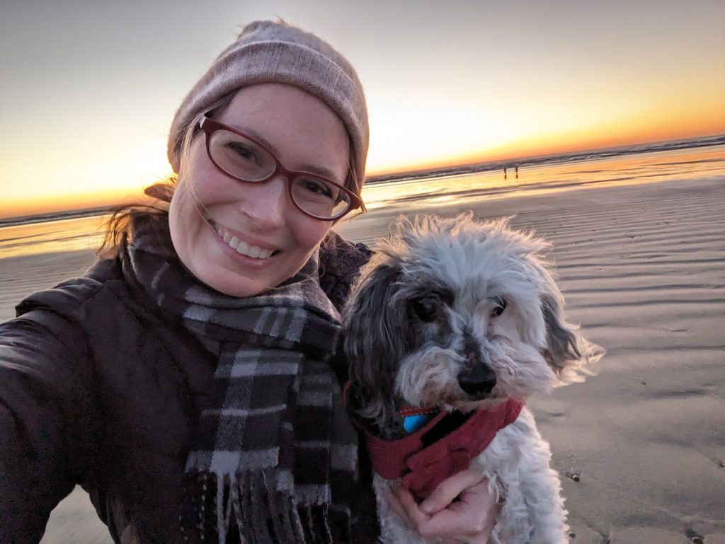 Amy in a coat, hat, and scarf holding a cute little dog in a red jacket, the sandy beach and sunset behind them